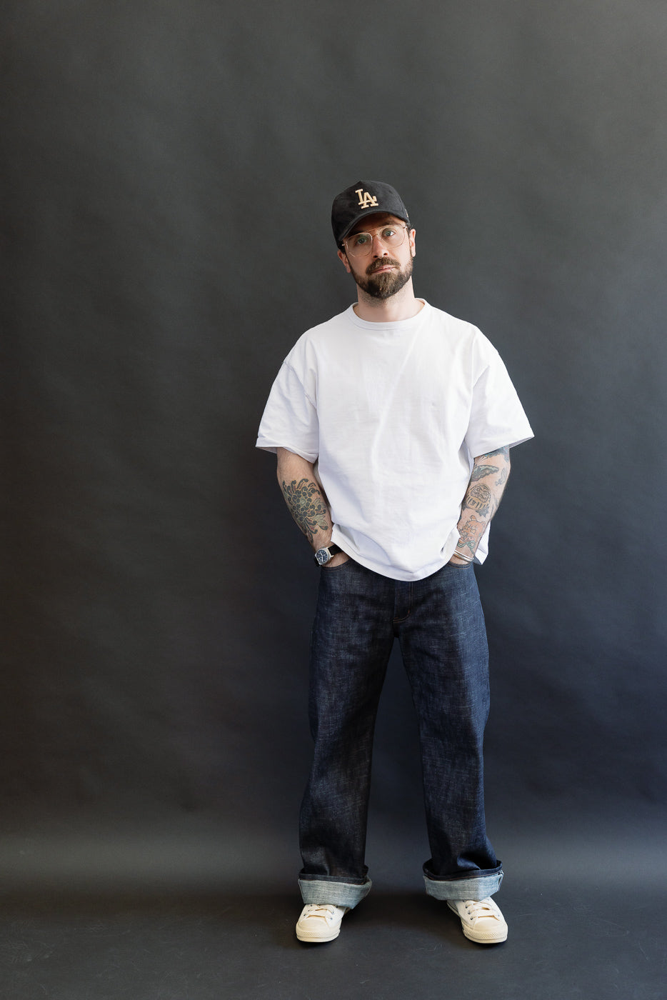 A man models the 12.5oz Greencast Stretch Selvedge - Strong Guy jeans, showcasing the relaxed fit with rolled cuffs and tattooed forearms, embodying Dant's premium menswear focus.