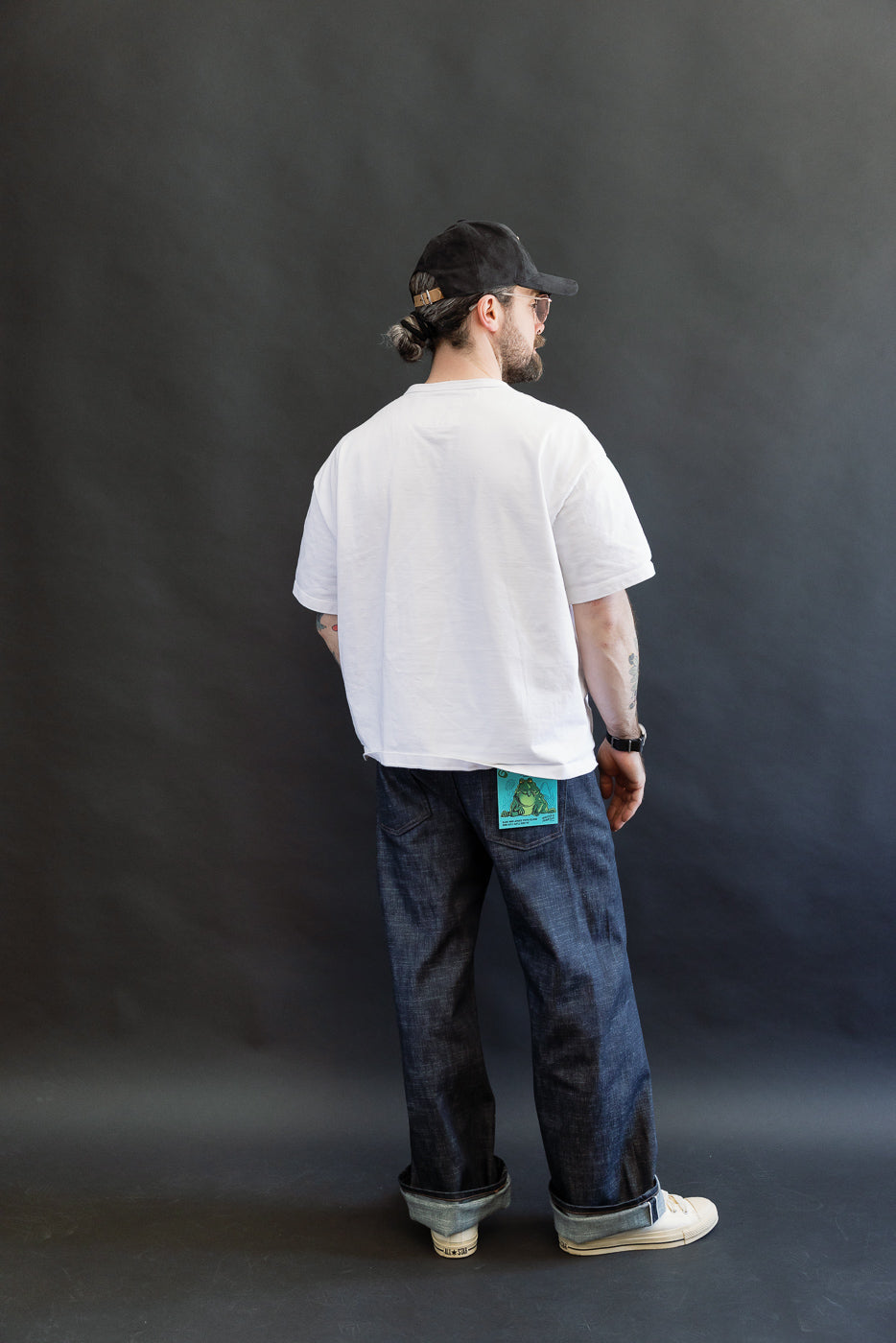 Man facing away, showcasing the 12.5oz Greencast Stretch Selvedge - Strong Guy jeans, cuffed with a colorful frog graphic. Paired with a white T-shirt, off-white sneakers, black cap, and sunglasses.