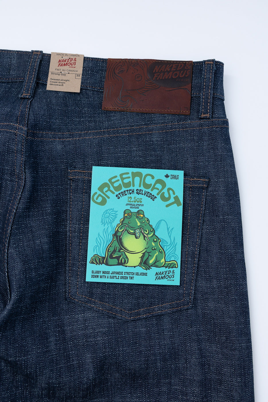 Rear view of Naked & Famous Denim's 12.5oz Greencast Stretch Selvedge - Strong Guy jeans, featuring a leather logo patch and a teal product card in the back pocket, highlighting Japanese stretch selvedge denim.