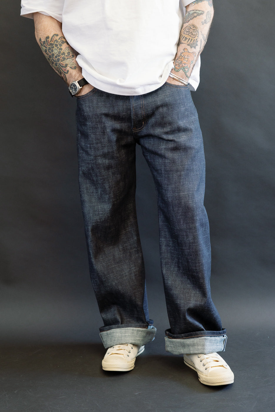Person stands wearing 12.5oz Greencast Stretch Selvedge - Strong Guy jeans, showcasing wide cuffs and tattooed forearms. Off-white sneakers and white T-shirt complete the casual look, emphasizing denim texture and style.