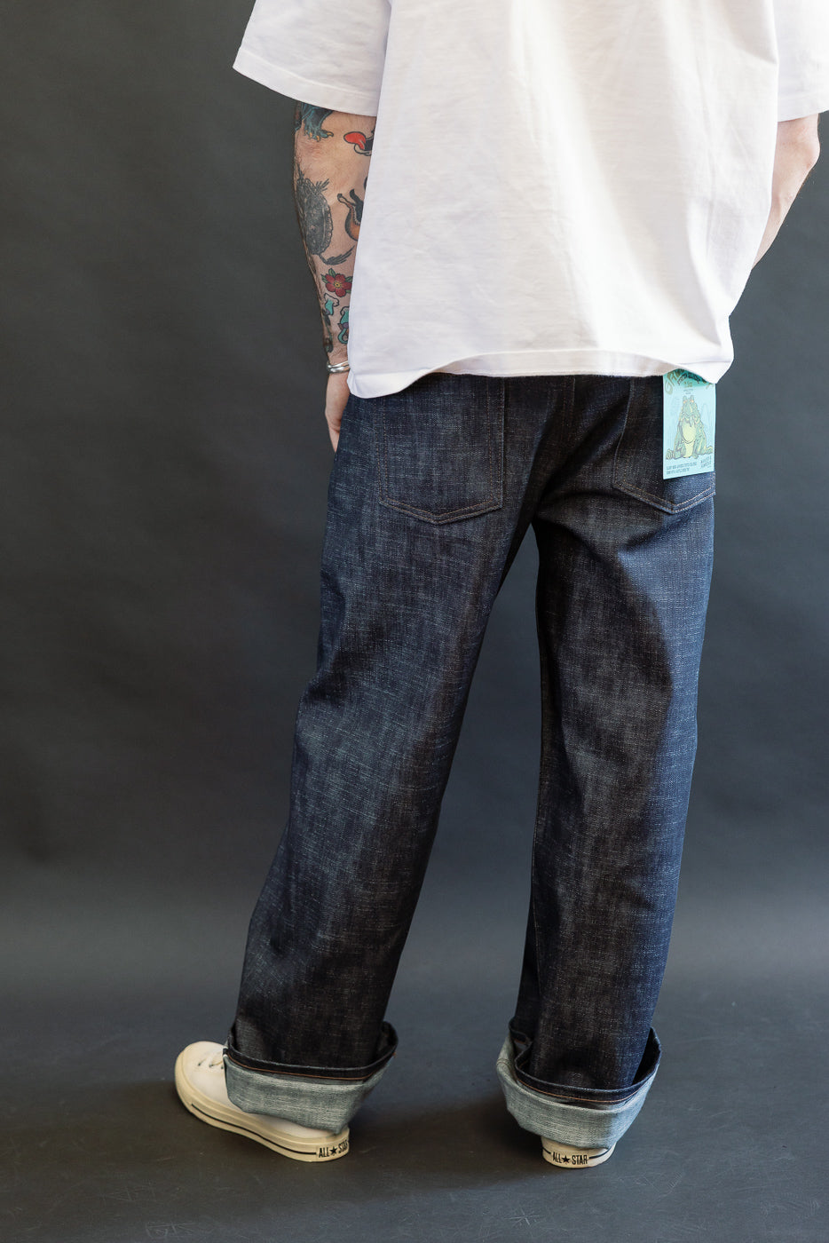 12.5oz - Greencast Stretch Selvedge - Strong Guy jeans featuring dark indigo straight-leg selvedge denim with a rolled cuff, paired with white low-top sneakers and a visible light blue tag in the back pocket.