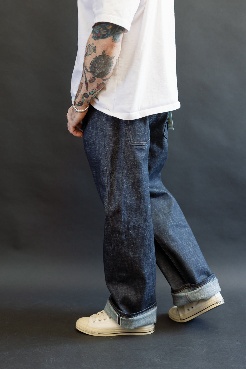 Person wearing 12.5oz Greencast Stretch Selvedge - Strong Guy jeans with cuffed hems, off-white canvas sneakers, a tattooed forearm, and a metallic bracelet, showcasing the denim's unique texture and style.