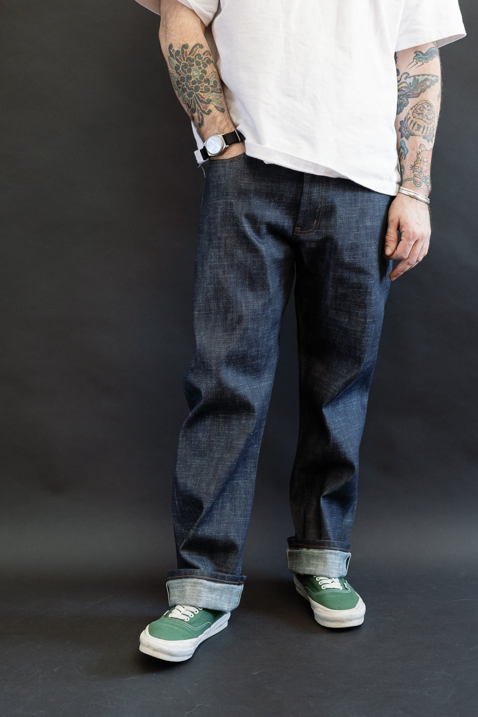 A person wears 12.5oz Greencast Stretch Selvedge - True Guy jeans, showcasing the straight-leg fit and wide cuff, paired with green canvas sneakers, tattoos visible on forearms, against a dark backdrop.