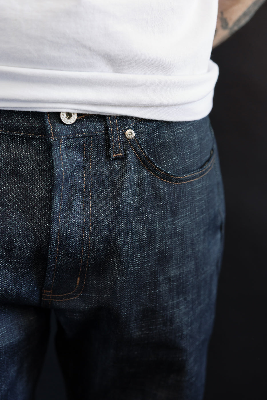 Close-up of the 12.5oz Greencast Stretch Selvedge True Guy jeans, showcasing the dark blue denim's button, belt loop, and pocket stitching. The fabric's texture highlights its unique slub construction and subtle green cast.