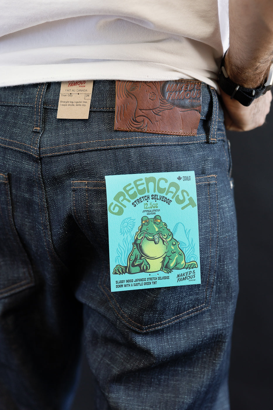Close-up of the lower back of a person wearing Naked & Famous 12.5oz Greencast Stretch Selvedge jeans, featuring a leather brand patch and teal promotional card, highlighting Japanese stretch selvedge denim.