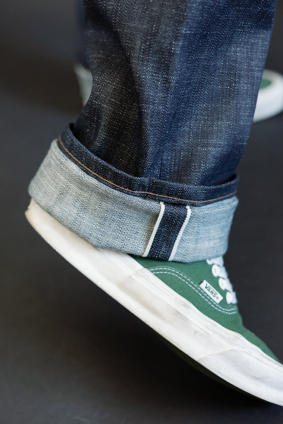 Close-up of the 12.5oz Greencast Stretch Selvedge - True Guy jeans, showcasing the rolled cuff with visible white-pink-white selvedge edge, paired with green canvas Vans sneaker, highlighting the jeans' texture and fit.