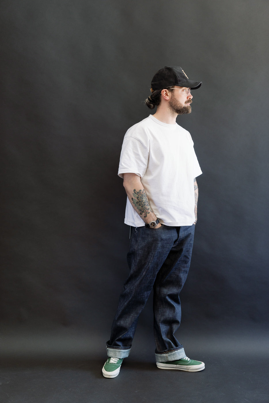 A 5'9 Caucasian male models the 12.5oz Greencast Stretch Selvedge - True Guy jeans, showcasing the unique indigo with a green cast and rolled cuffs, paired with a white T-shirt and green sneakers.