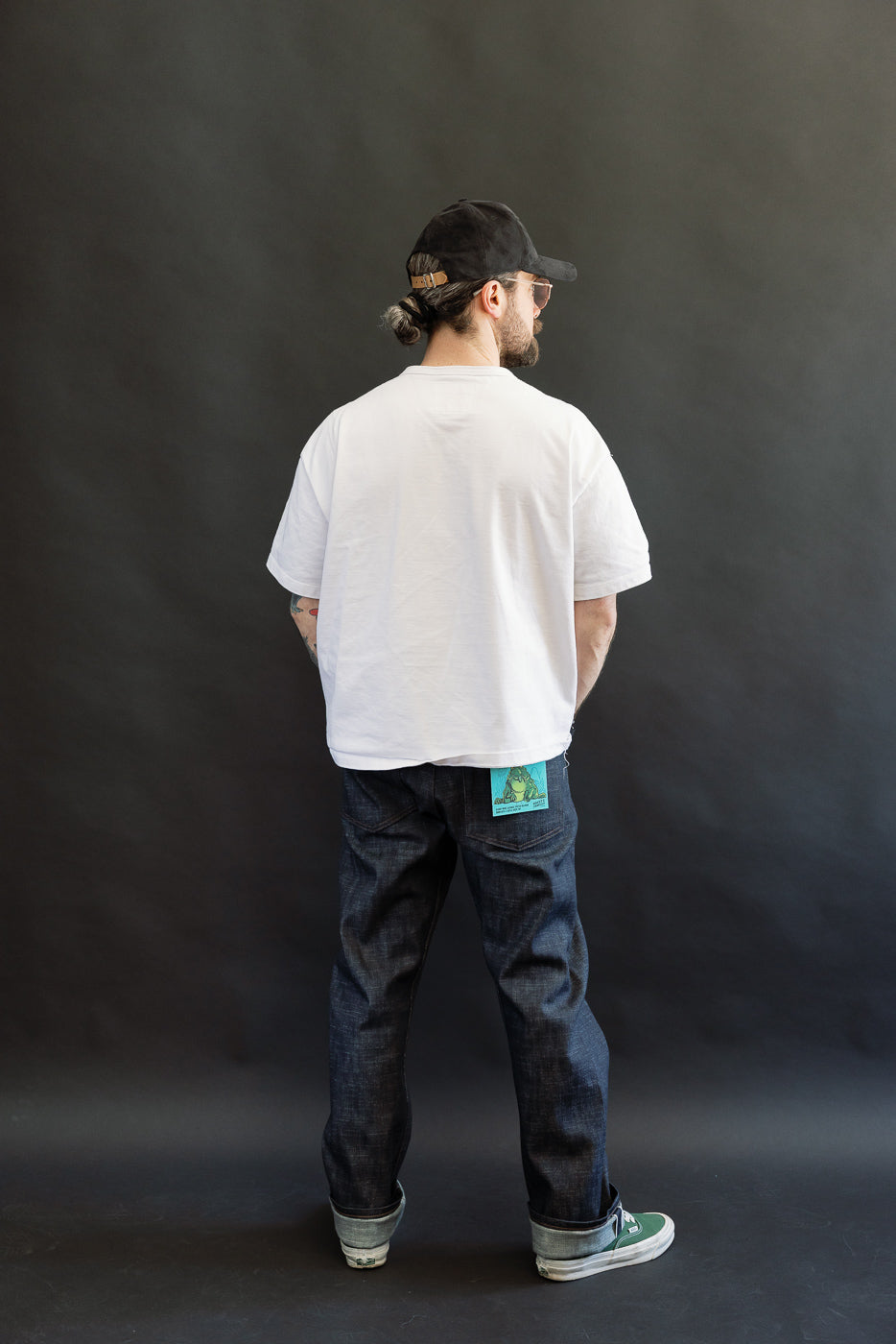 A man stands with his back to the camera, showcasing the 12.5oz Greencast Stretch Selvedge - True Guy jeans, emphasizing their straight fit and subtle turquoise detail, styled casually with sneakers and a cap.
