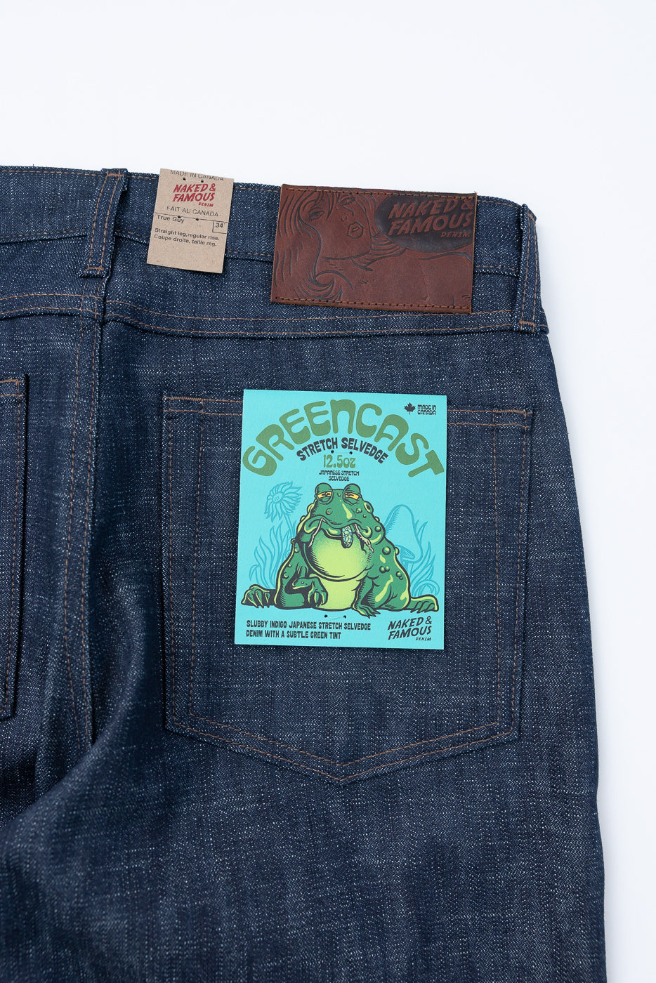 Back view of Naked & Famous 12.5oz - Greencast Stretch Selvedge - True Guy jeans, showcasing waxed leather brand patch and green-blue hang-tag with frog illustration, emphasizing Japanese selvedge denim's unique texture and comfort.