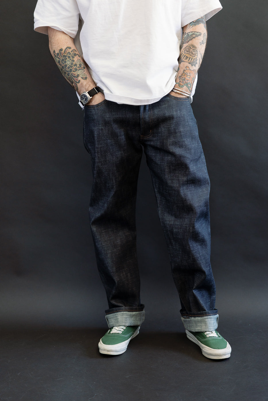 12.5oz Greencast Stretch Selvedge - True Guy jeans showcased with rolled cuffs, revealing intricate tattoos on forearms, paired with green canvas sneakers. The jeans' depth and texture are accentuated by the selvedge's unique green cast.