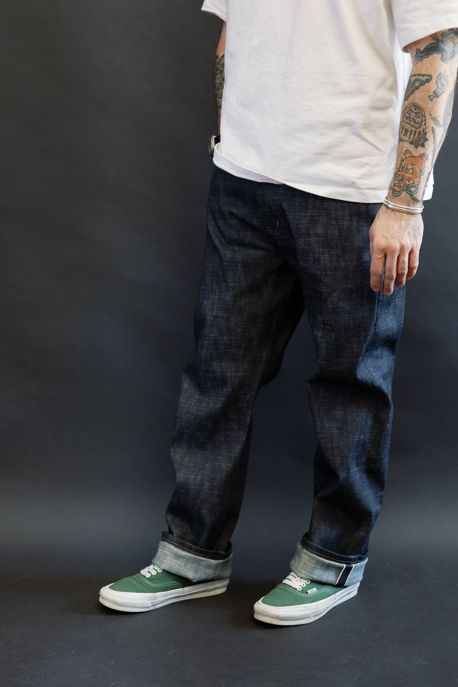 Person wearing 12.5oz Greencast Stretch Selvedge - True Guy jeans with rolled cuffs, displaying tattooed forearms, bracelets, and green canvas sneakers. The denim features slub construction and a subtle green cast.