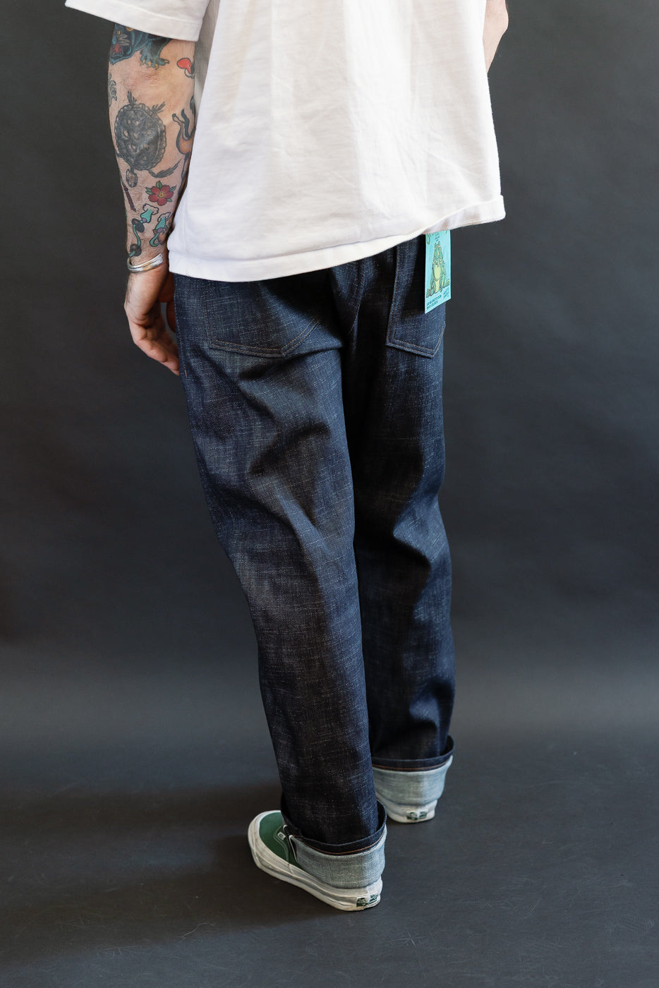 Person wearing 12.5oz - Greencast Stretch Selvedge - True Guy jeans with rolled cuffs, revealing selvedge edge; paired with slip-on skate shoes and visible tattoos, showcasing Dant's premium denim craftsmanship.