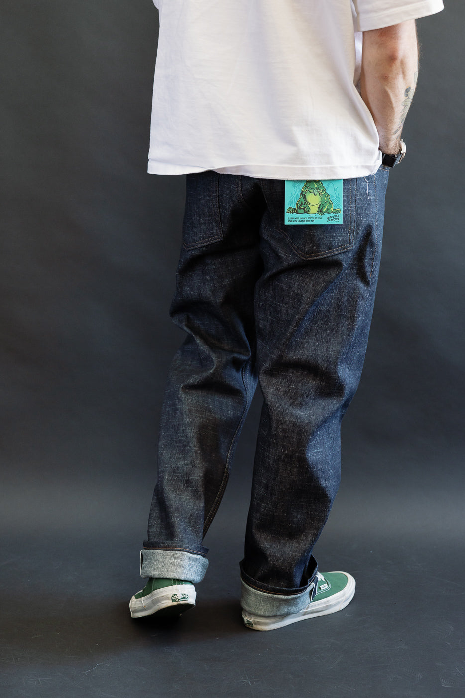 12.5oz Greencast Stretch Selvedge - True Guy jeans feature dark denim with rolled cuffs, accented by a turquoise-green card in the back pocket. Paired with white slip-on sneakers and a white T-shirt.