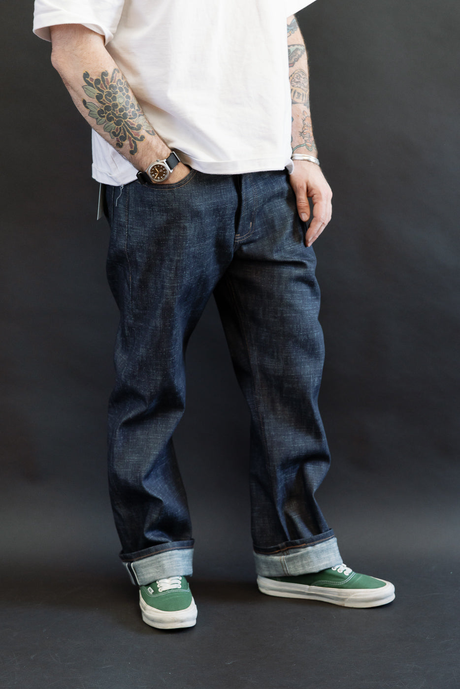12.5oz - Greencast Stretch Selvedge - True Guy jeans with rolled cuffs, revealing lighter inside. Styled with green canvas sneakers, forearm tattoos, and casual hand in pocket, showcasing premium denim's texture and fit.