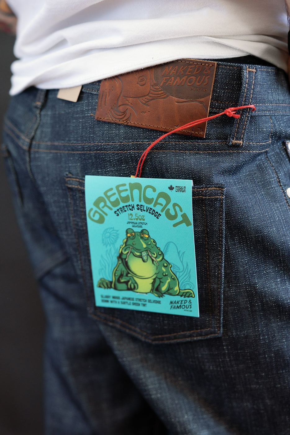 Close-up of the 12.5oz - Greencast Stretch Selvedge - Weird Guy jeans waistband, featuring a brown leather patch and teal-green hang-tag, showcasing the denim's selvage-style stitching.