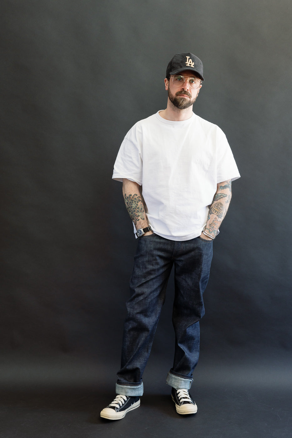Man wearing 12.5oz Greencast Stretch Selvedge - Weird Guy jeans, showcasing tattoos, white T-shirt, and black sneakers. The denim's slub texture and tailored fit emphasize comfort and style in a relaxed pose.