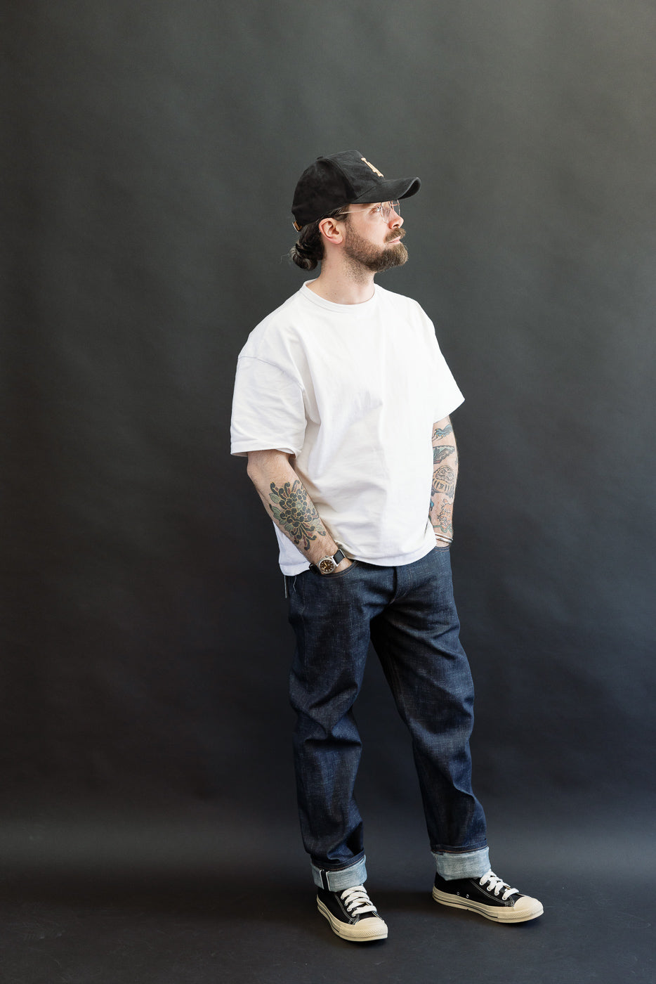 Man wearing 12.5oz Greencast Stretch Selvedge - Weird Guy denim, featuring a relaxed fit with rolled cuffs, styled with a white T-shirt and sneakers. He stands with hands in pockets, showcasing colorful arm tattoos.