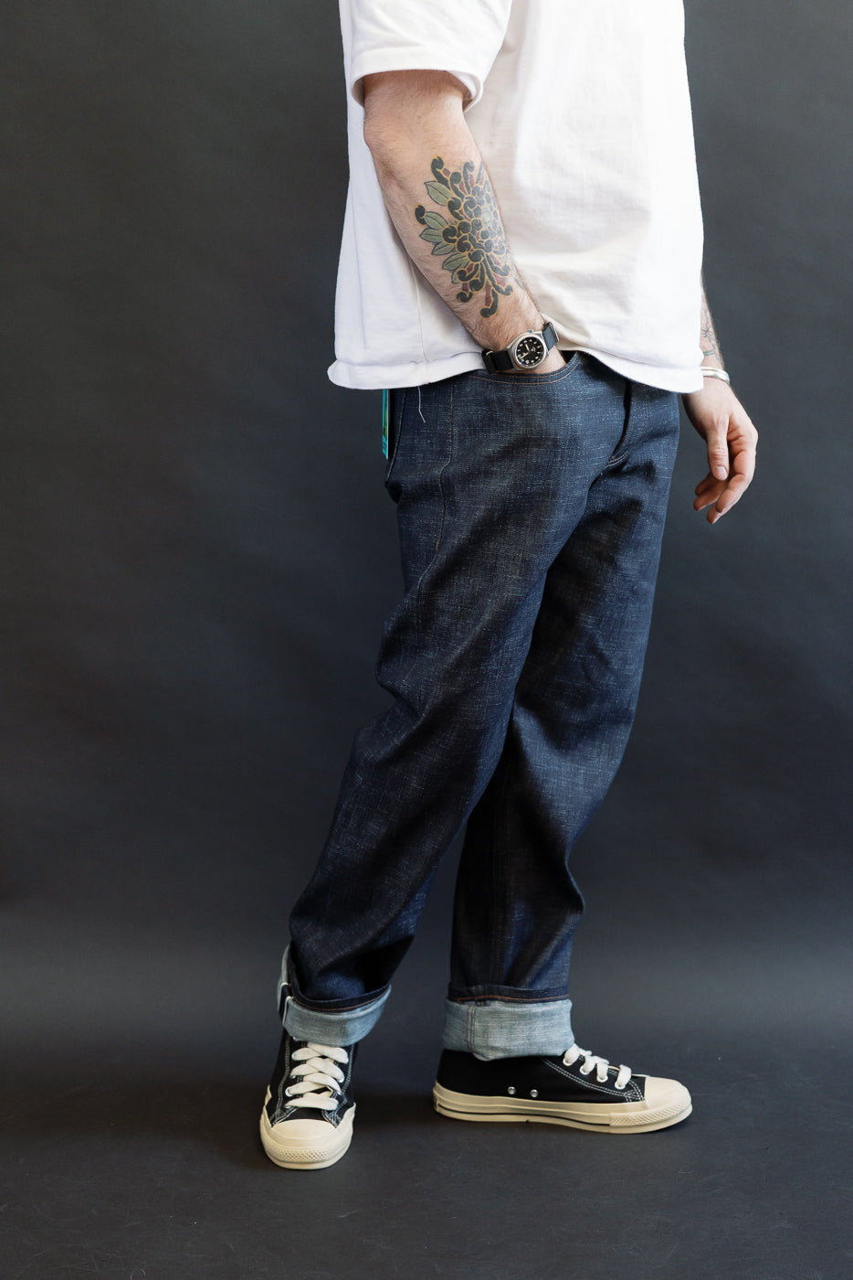 Person wearing Naked & Famous 12.5oz Greencast Stretch Selvedge Weird Guy jeans, showcasing dark indigo denim with rolled cuffs, paired with black high-top sneakers, a white T-shirt, and a visible floral tattoo.