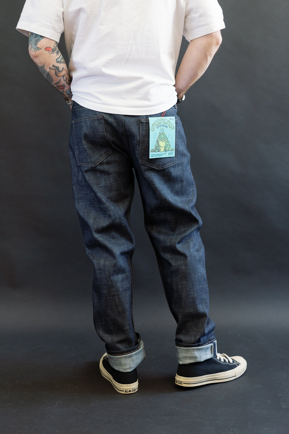 The 12.5oz Greencast Stretch Selvedge - Weird Guy jeans, featuring cuffed dark denim with a mint-green GREENCAST tag, are paired with a white T-shirt and black canvas sneakers.