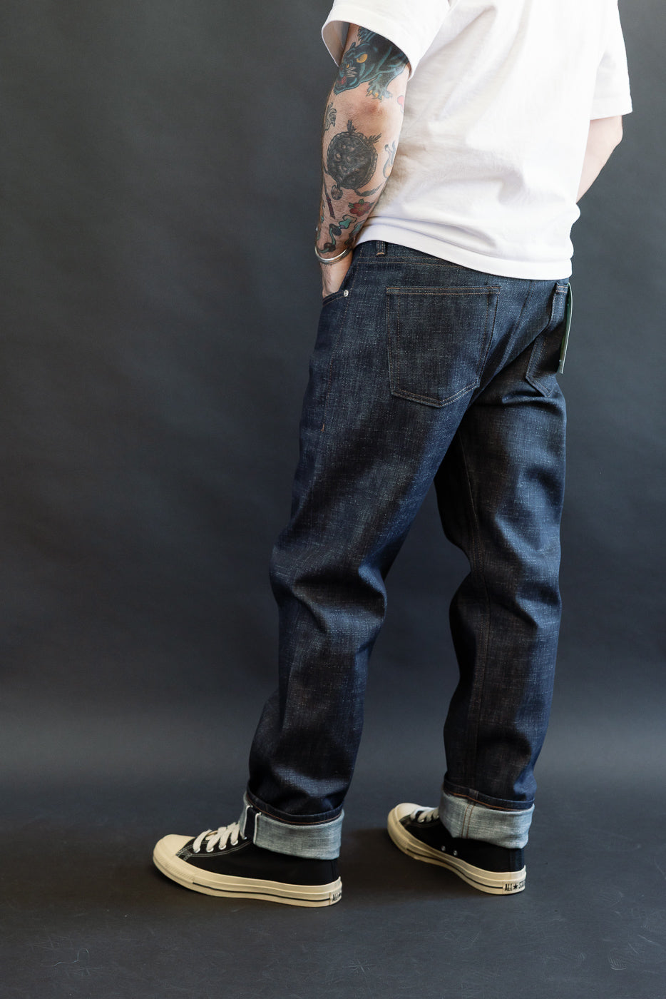 The lower half of a person showcases the 12.5oz Greencast Stretch Selvedge - Weird Guy jeans, highlighting the rolled cuffs and black low-top canvas sneakers, with one tattooed forearm visible against a dark-gray background.