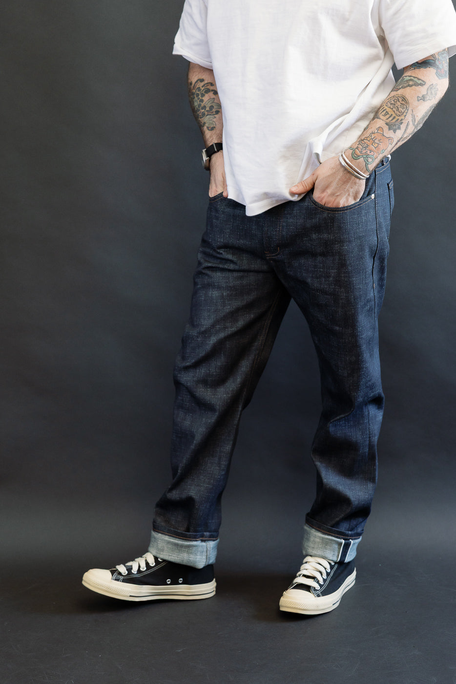 12.5oz - Greencast Stretch Selvedge - Weird Guy jeans, dark indigo with wide cuffs, worn with casual hands-in-pockets stance, showcasing tattoos. Styled with a plain white T-shirt and black canvas sneakers.