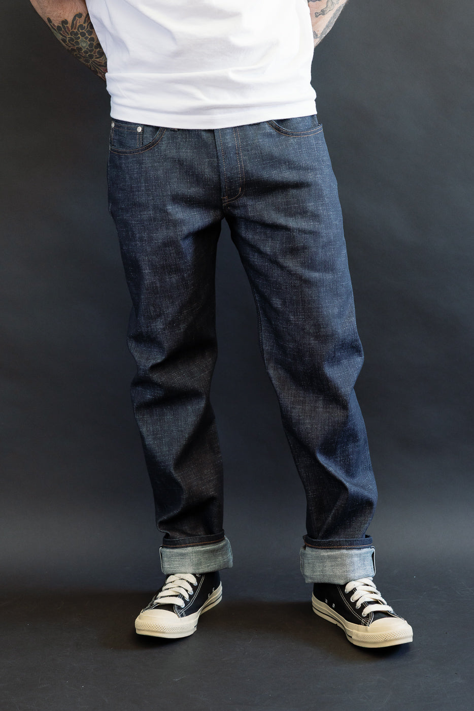 Lower half of a person wearing 12.5oz Greencast Stretch Selvedge - Weird Guy jeans, showcasing rolled cuffs and black canvas sneakers against a dark background. Tattooed forearms visible above the waistline.