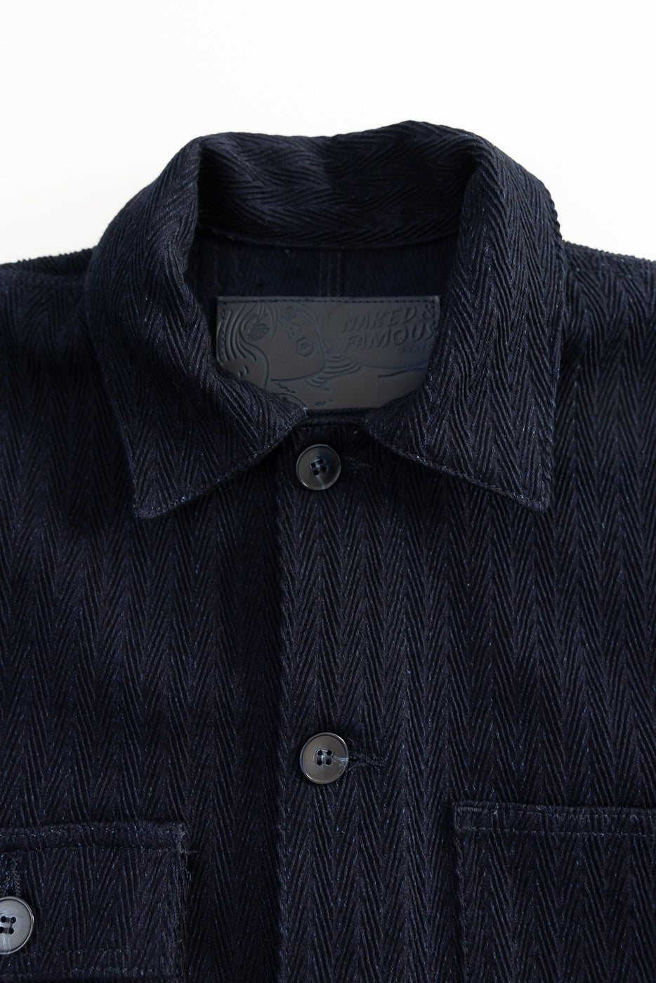 Close-up front of Naked & Famous indigo chore coat collar, herringbone textured selvedge fabric, buttons, and leather brand patch at Dant.