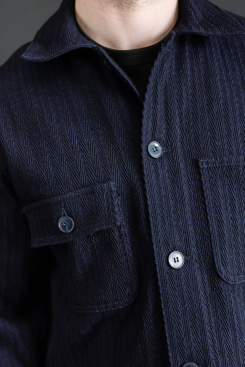 Close-up of front chest and collar of Naked & Famous indigo chore coat on 5'9 male model, showing herringbone fabric texture, chest pocket, and button details.