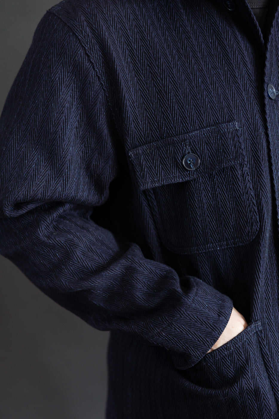 Close-up of front left chest and midsection of indigo herringbone chore coat on male model, showing buttoned pocket, textured fabric, and cuff detail, Dant contemporary casual apparel.
