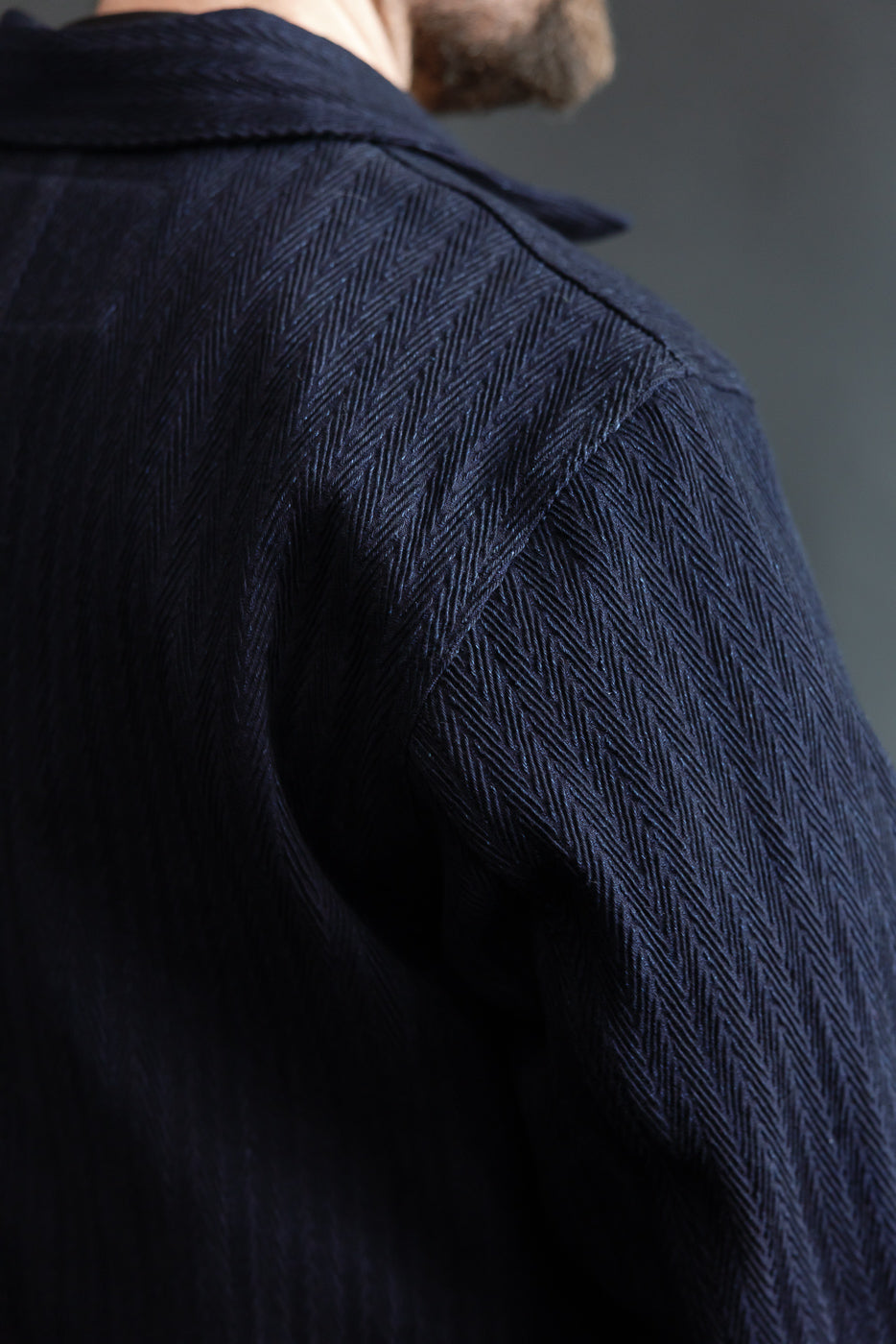 Close-up of male model’s upper back and shoulder in Naked & Famous indigo chore coat, showing textured herringbone selvedge fabric detail, back view. Model is 5'9