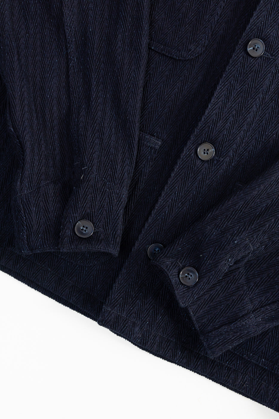 Close-up of front lower panel on Naked & Famous 12oz Indigo Sugiaya chore coat, showing herringbone selvedge fabric, front buttons, and sleeve cuffs. No model visible.