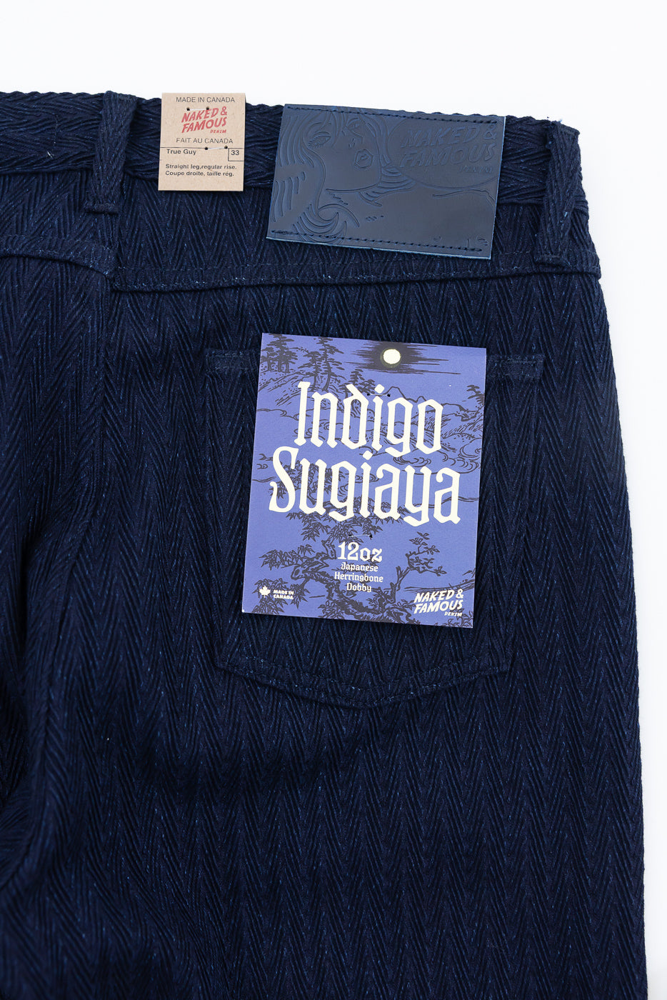 Close-up of Naked & Famous Indigo Sugiaya jeans back pocket with herringbone texture, leather patch, and product tags visible.