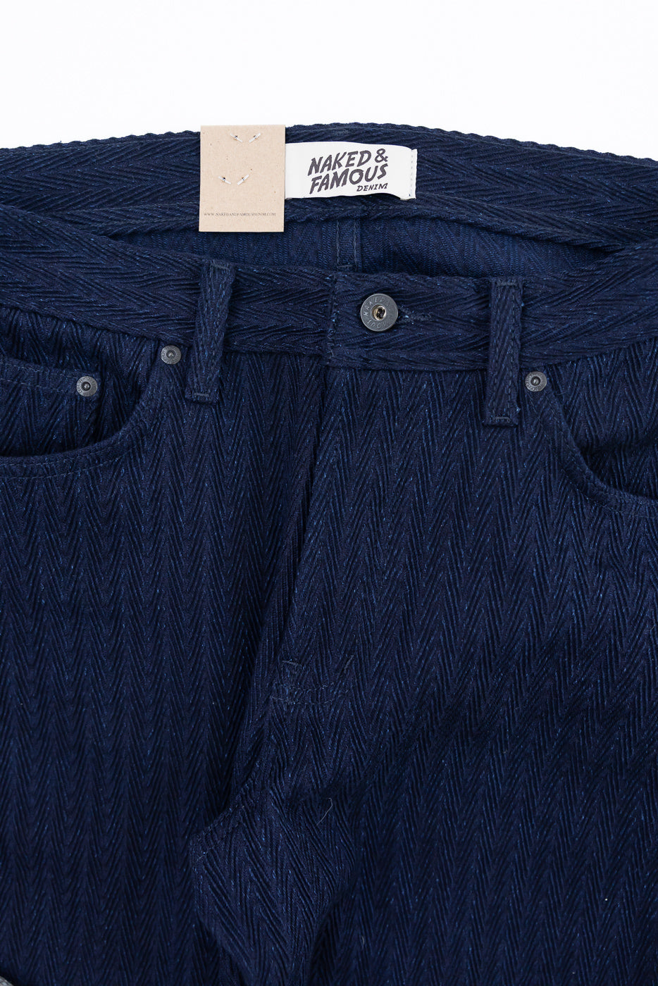 Close-up front of Naked & Famous indigo selvedge jeans showing button, waistband, and herringbone fabric texture.
