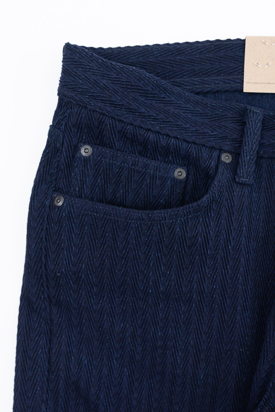 Close-up of front left waist and pocket on Naked & Famous 12oz indigo selvedge jeans, showing herringbone fabric and rivet details.