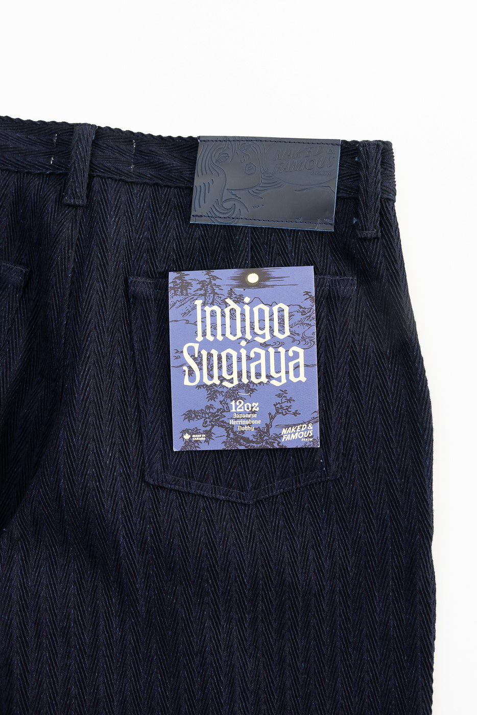 Close-up of Naked & Famous Indigo Sugiaya pleated trouser back waist with herringbone texture, rear pocket, leather patch, and product tag, Dant contemporary casual apparel.