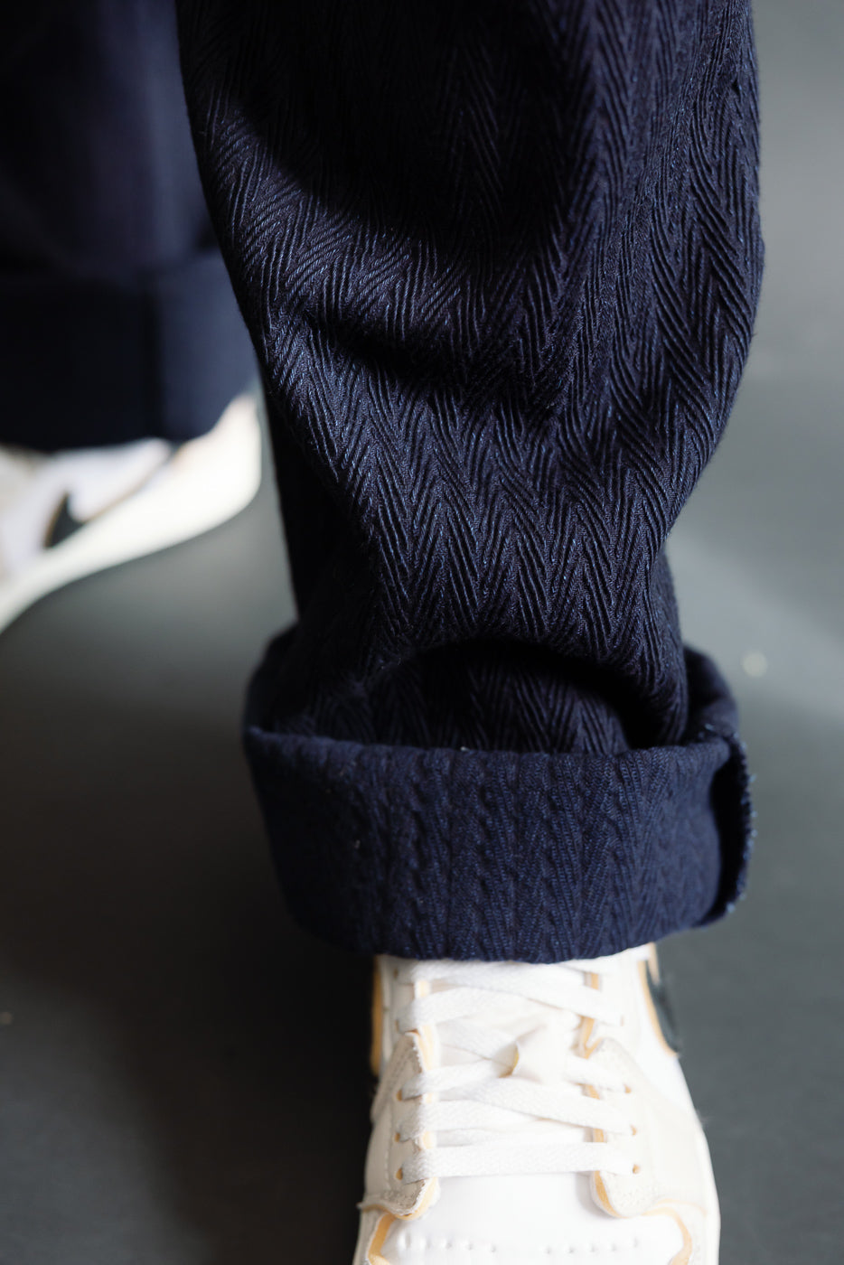 Close-up of cuffed indigo pleated trouser leg, showing herringbone texture and sneaker on male model, Dant contemporary apparel.