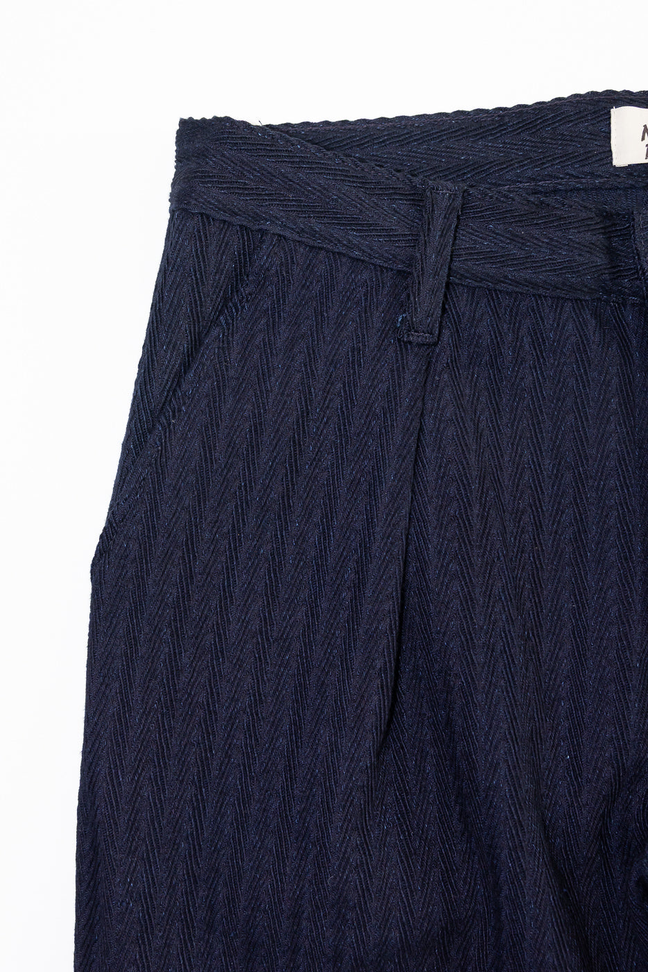 Close-up front view of Naked & Famous indigo pleated trouser, showing textured herringbone fabric, waistband, belt loop, and pocket detail.