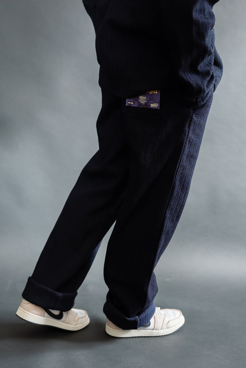 Male model 5'9” 165lbs wearing Naked & Famous indigo pleated trouser, right side view, visible rear pocket tag, full body.