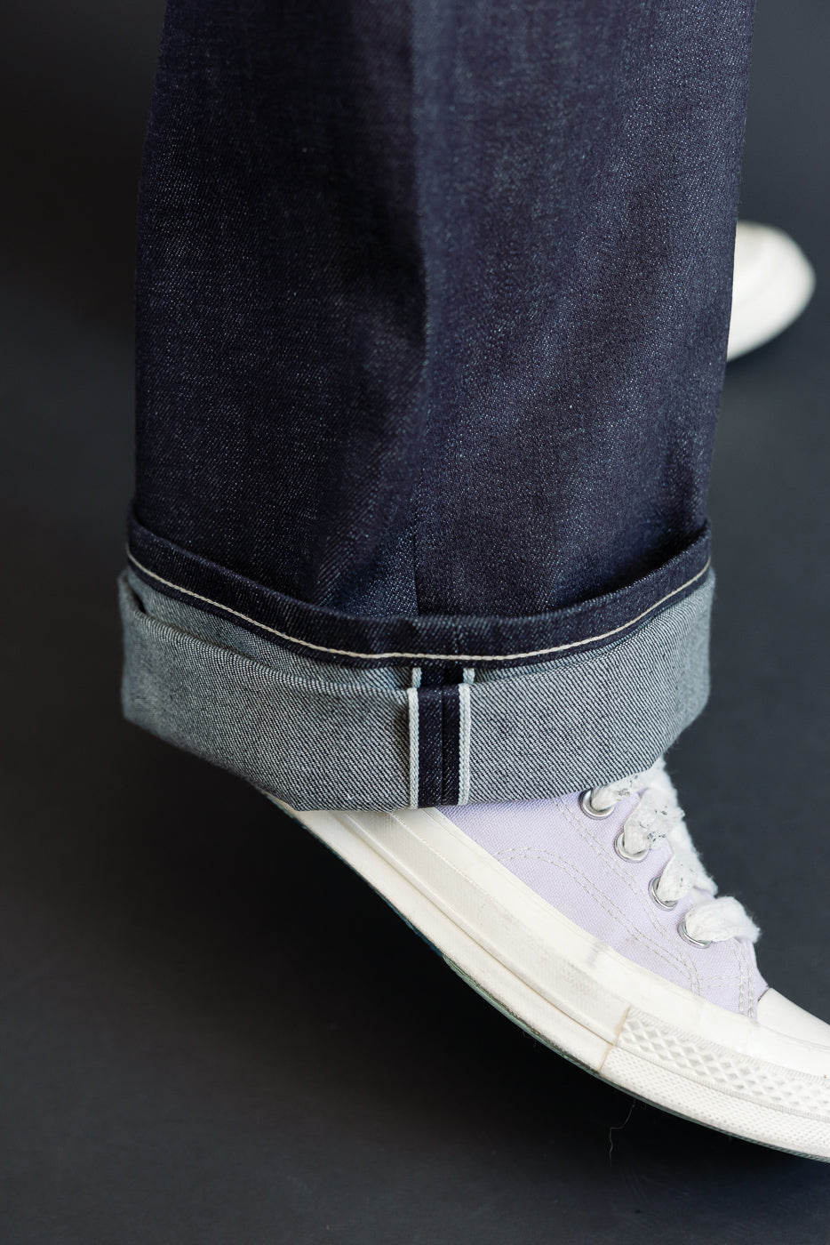 Close-up of a male model's lower leg showing a selvedge denim cuff and white sneakers.