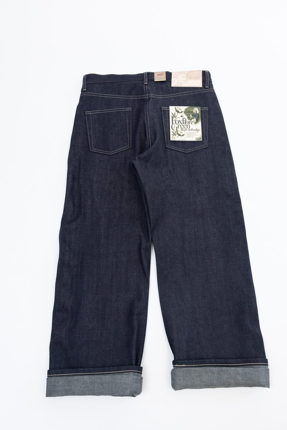 Back view flat lay of dark indigo selvedge denim jeans with rear patch pockets and cuffed hems.