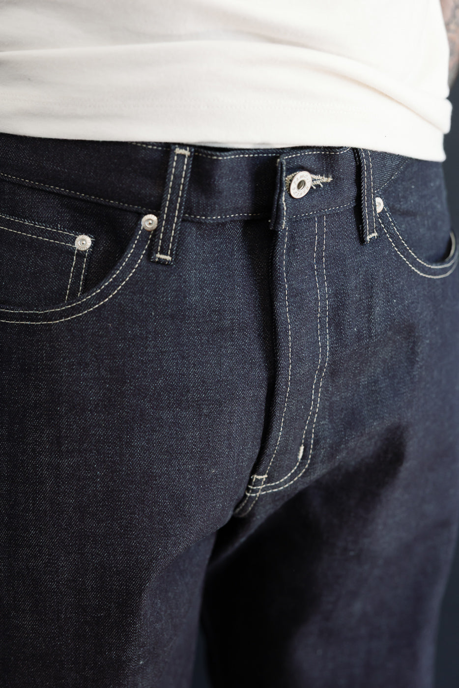 Close-up front view of dark indigo selvedge denim jeans on a male model, showing waist and pocket details.