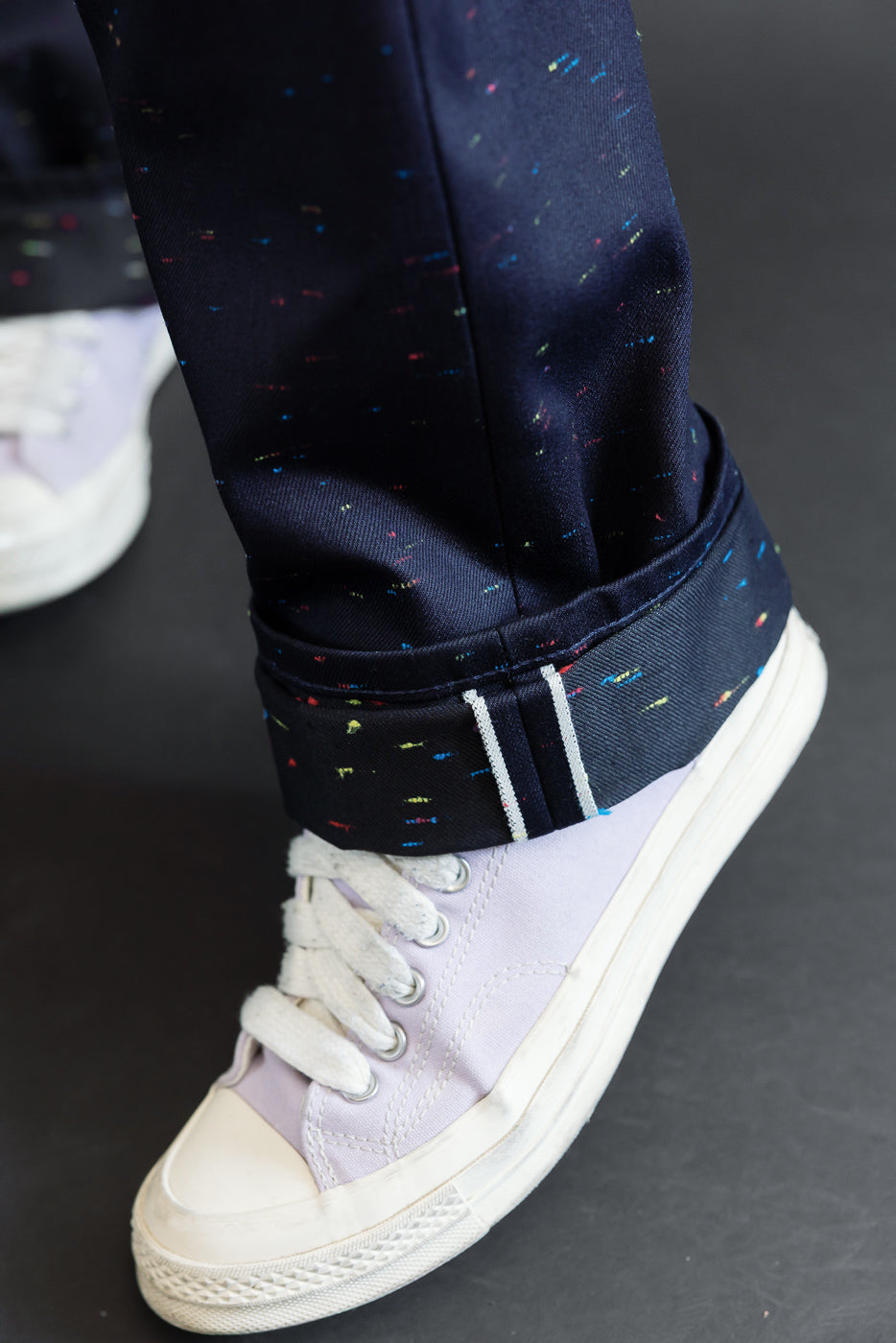 Close-up of the cuffed hem of navy blue speckled trousers paired with light purple canvas sneakers.