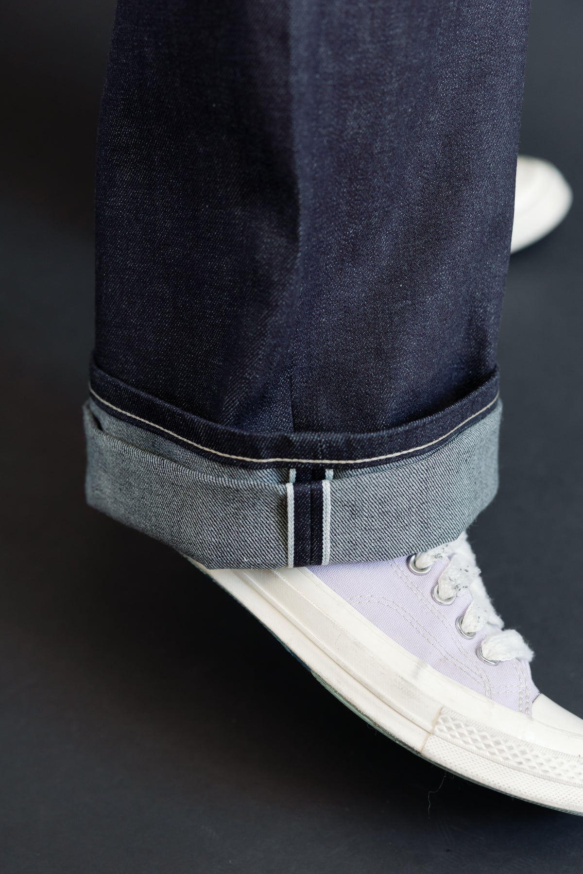 Close-up of a male model's lower leg showing the cuffed hem of dark selvedge denim jeans.
