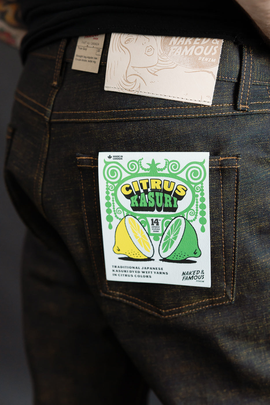 Close-up of back side of men's selvedge jeans on male model, 5'9, 165lbs, athletic build, showing denim texture, rear pocket, branded leather patch, and Citrus Kasuri paper tag.