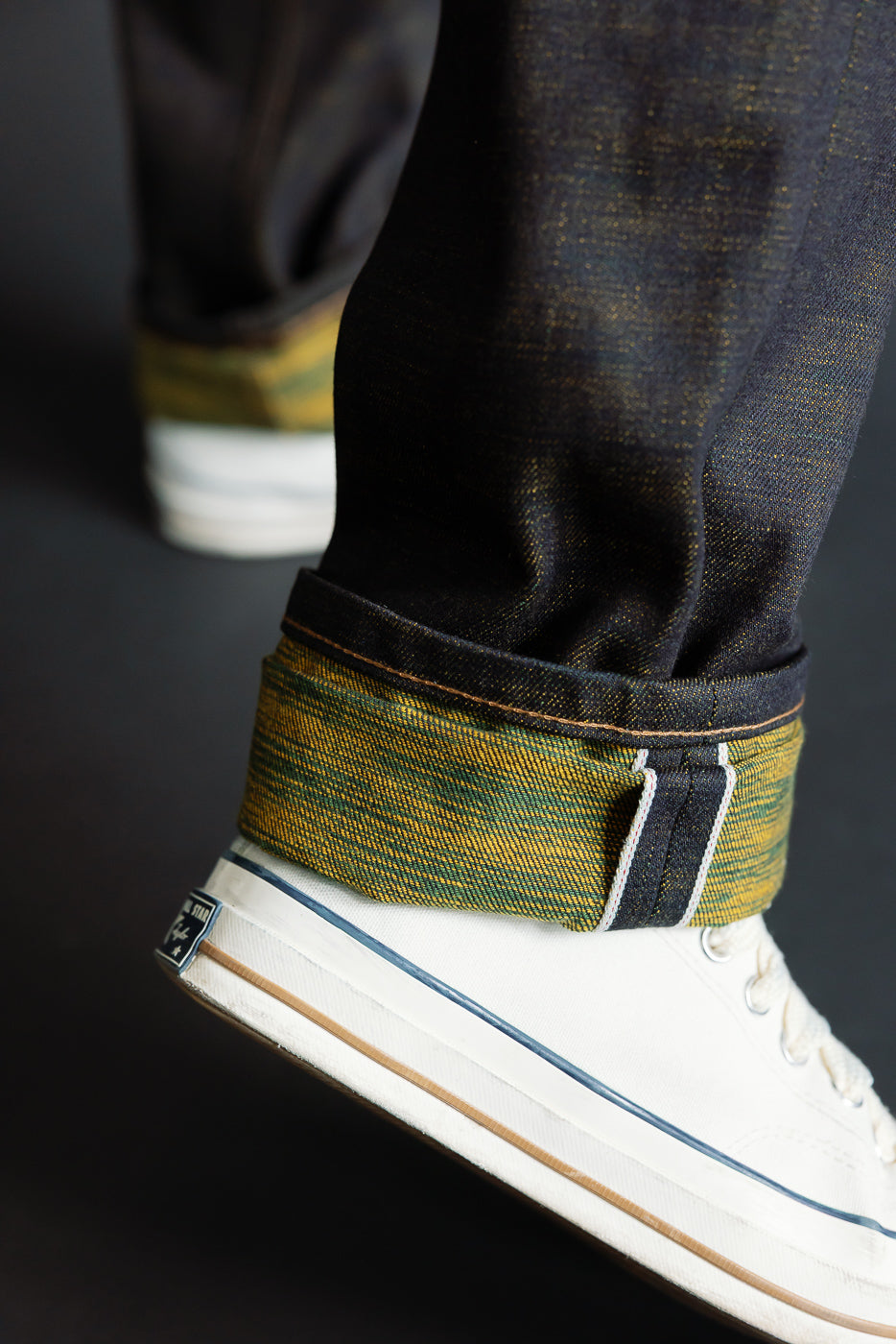 Male model close-up of cuffed selvedge denim jeans hem, side view, highlighting yellow-green weft detail above white sneakers. Model is 5'9
