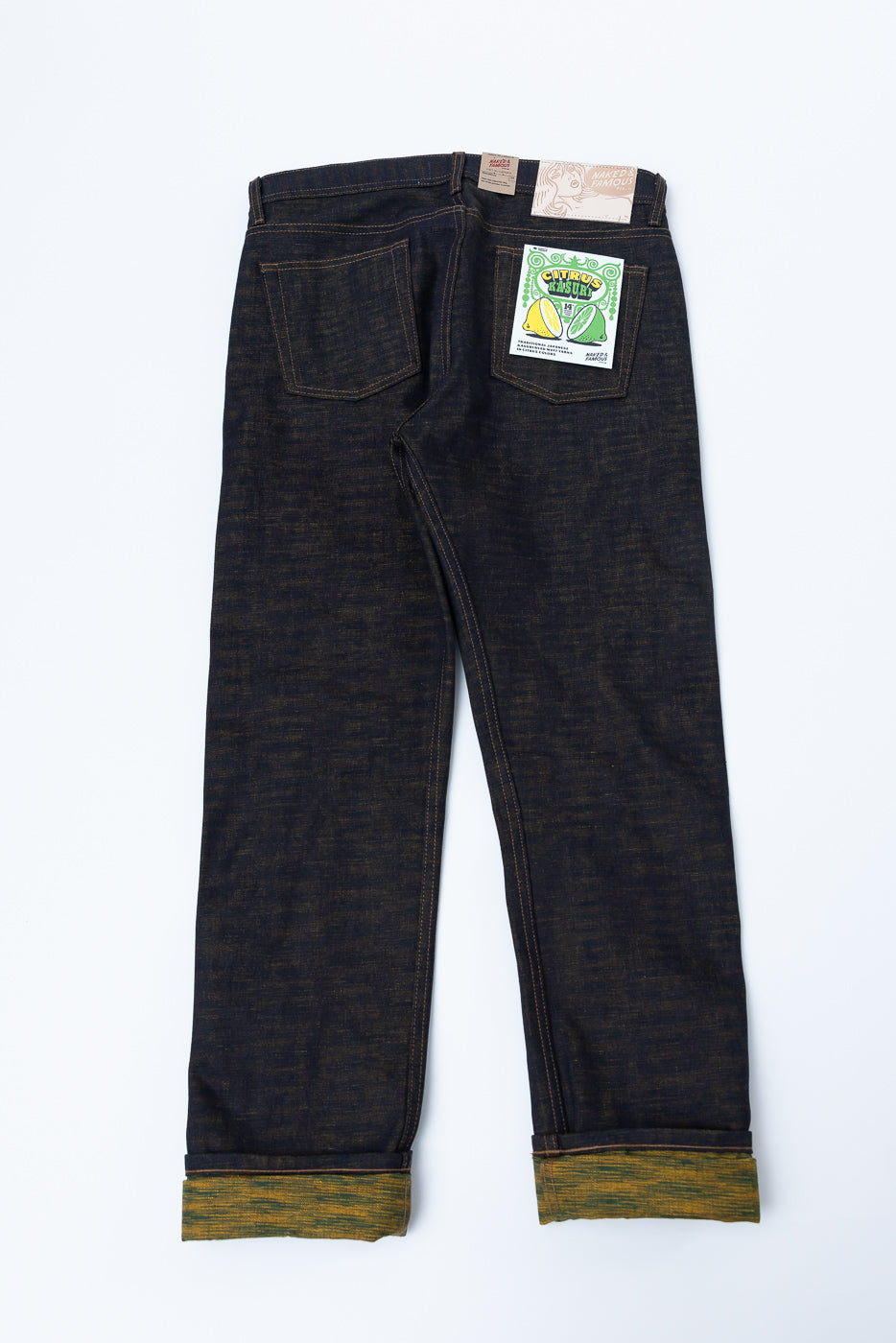 Flat lay of dark indigo selvedge jeans, back view, showing rear pockets, leather patch, and colorful tag