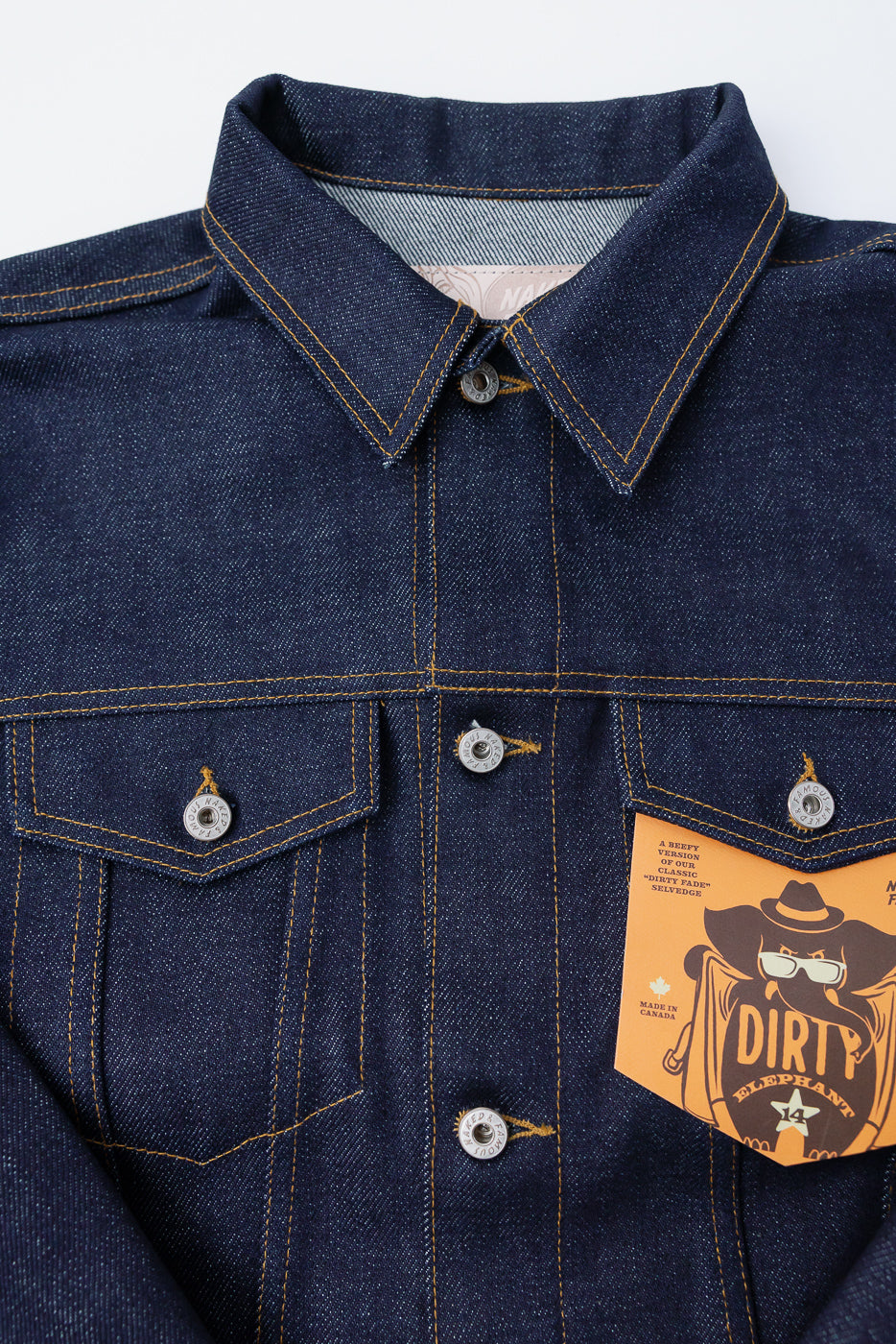 Close-up front view of Naked & Famous 20oz Dirty Elephant selvedge denim jacket, showing chest pockets, metal buttons, pointed collar, and visible yellow stitching detail.