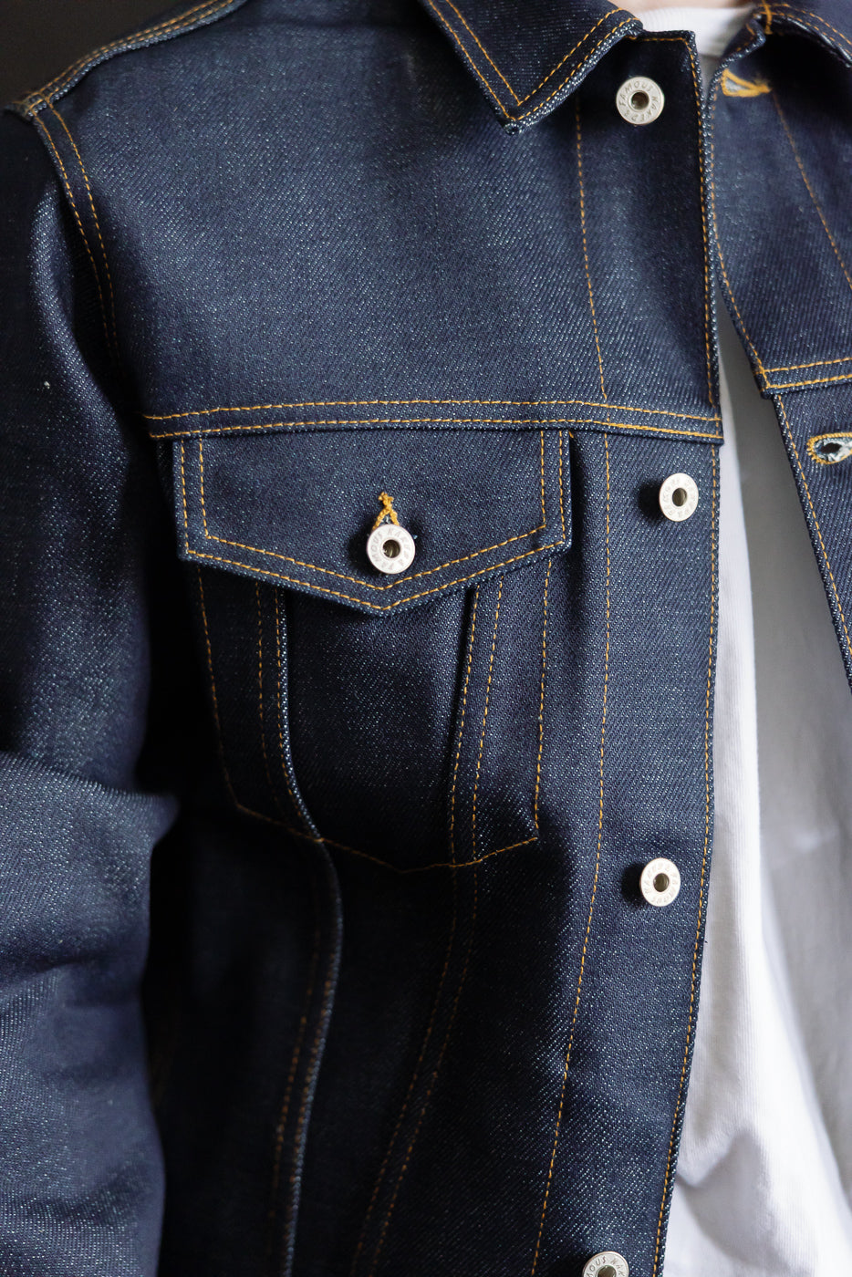 Close-up front detail of selvedge denim jacket chest pocket, buttons, and contrast stitching.