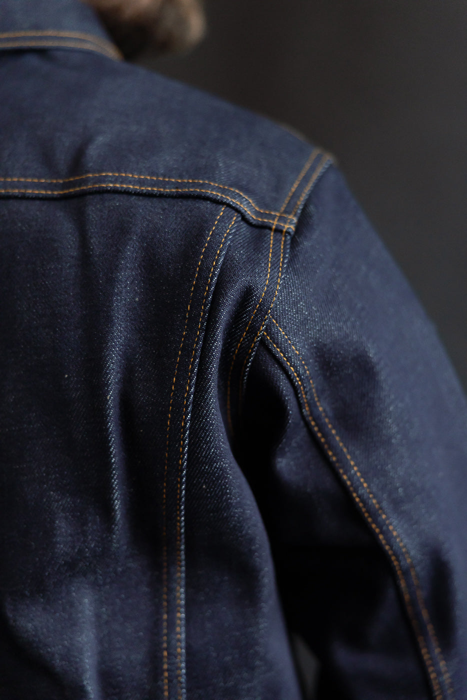 Close-up of men's selvedge denim jacket shoulder and back yoke, highlighting fabric texture and stitching detail.