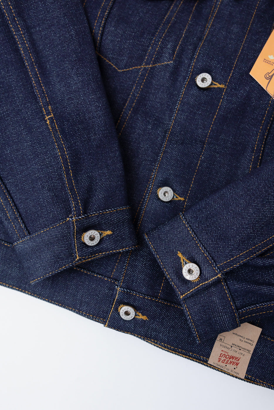 Close-up of front hem and cuff details on Naked & Famous 20oz Dirty Elephant selvedge denim jacket, highlighting metal buttons, orange stitching, and brand tag.